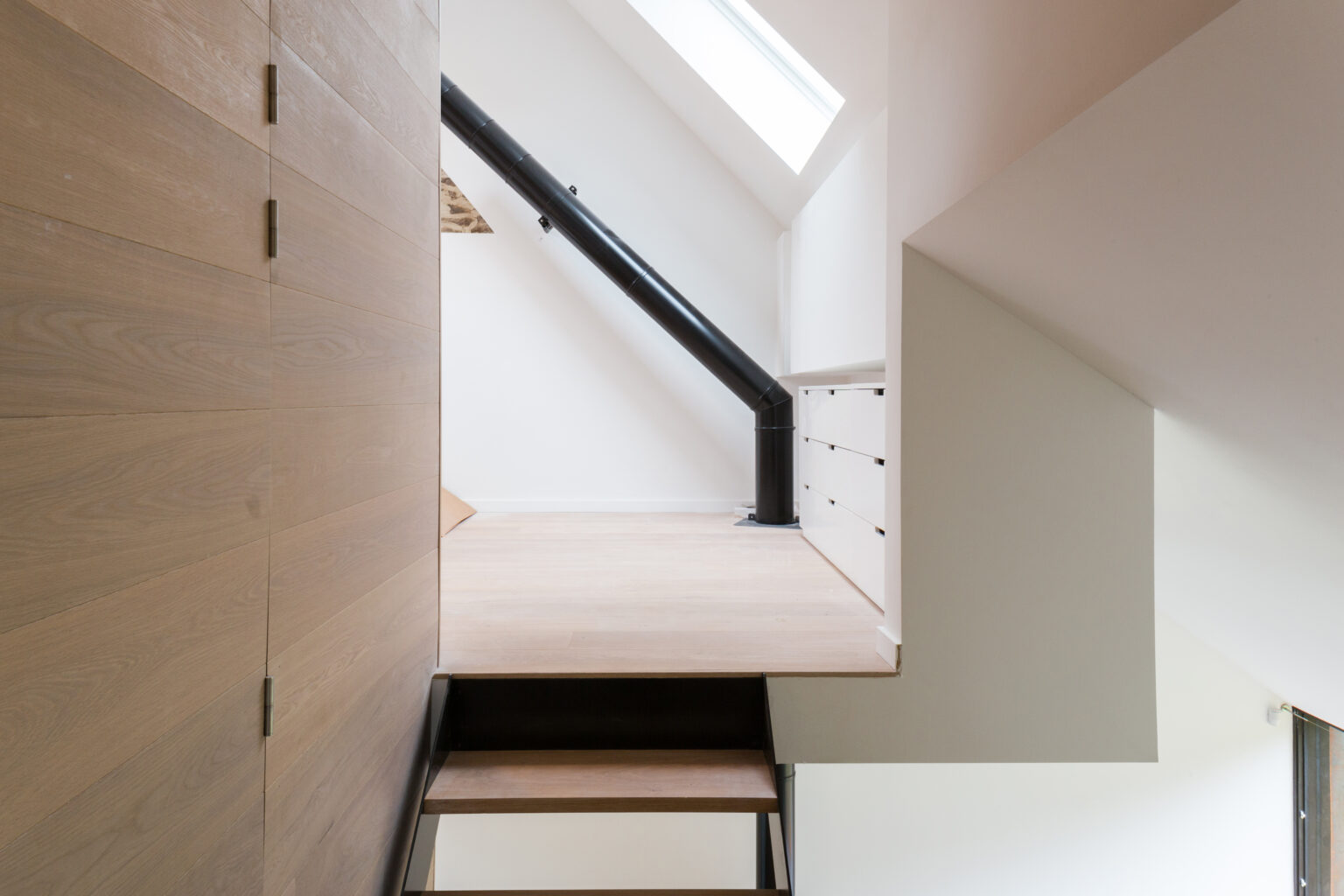 Escalier en bois clair et garde-corps métallique noir sous la charpente, rénovation à Lustin